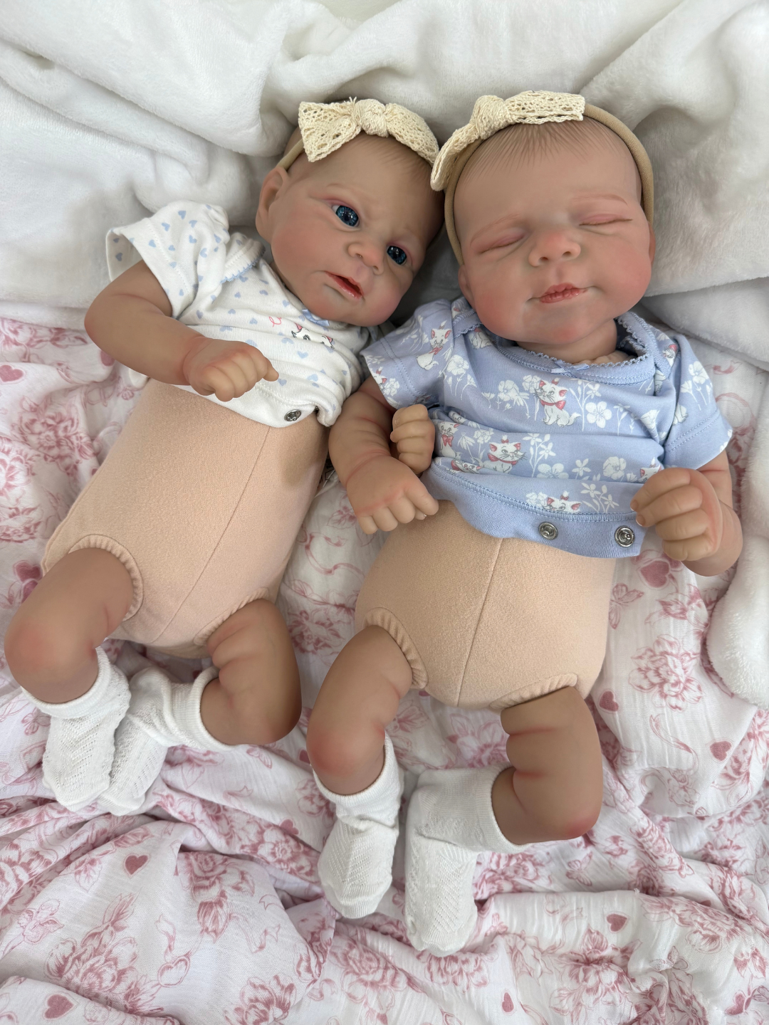 Two realistic reborn baby dolls lying side by side on a pink patterned blanket with a white throw. The left doll wears a white top with small blue floral prints, beige pants, white socks, and a cream bow headband.