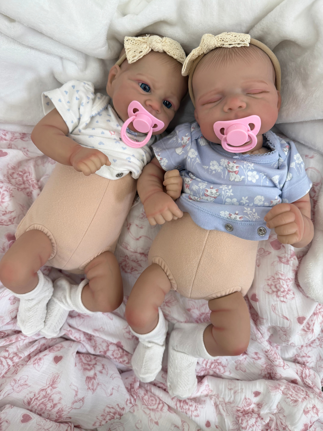 Two realistic baby dolls lying side by side on a pink and white patterned blanket. One doll is awake wearing a white floral top, peach pants, white socks, and a cream bow headband with a pink pacifier.