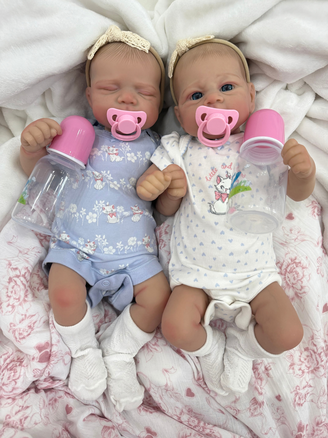 Two realistic baby dolls lying side by side on a white blanket, each holding a clear bottle with a pink cap and wearing pacifiers. One doll wears a light blue floral onesie and headband, and the other wears a white onesie with a bunny print and headband.