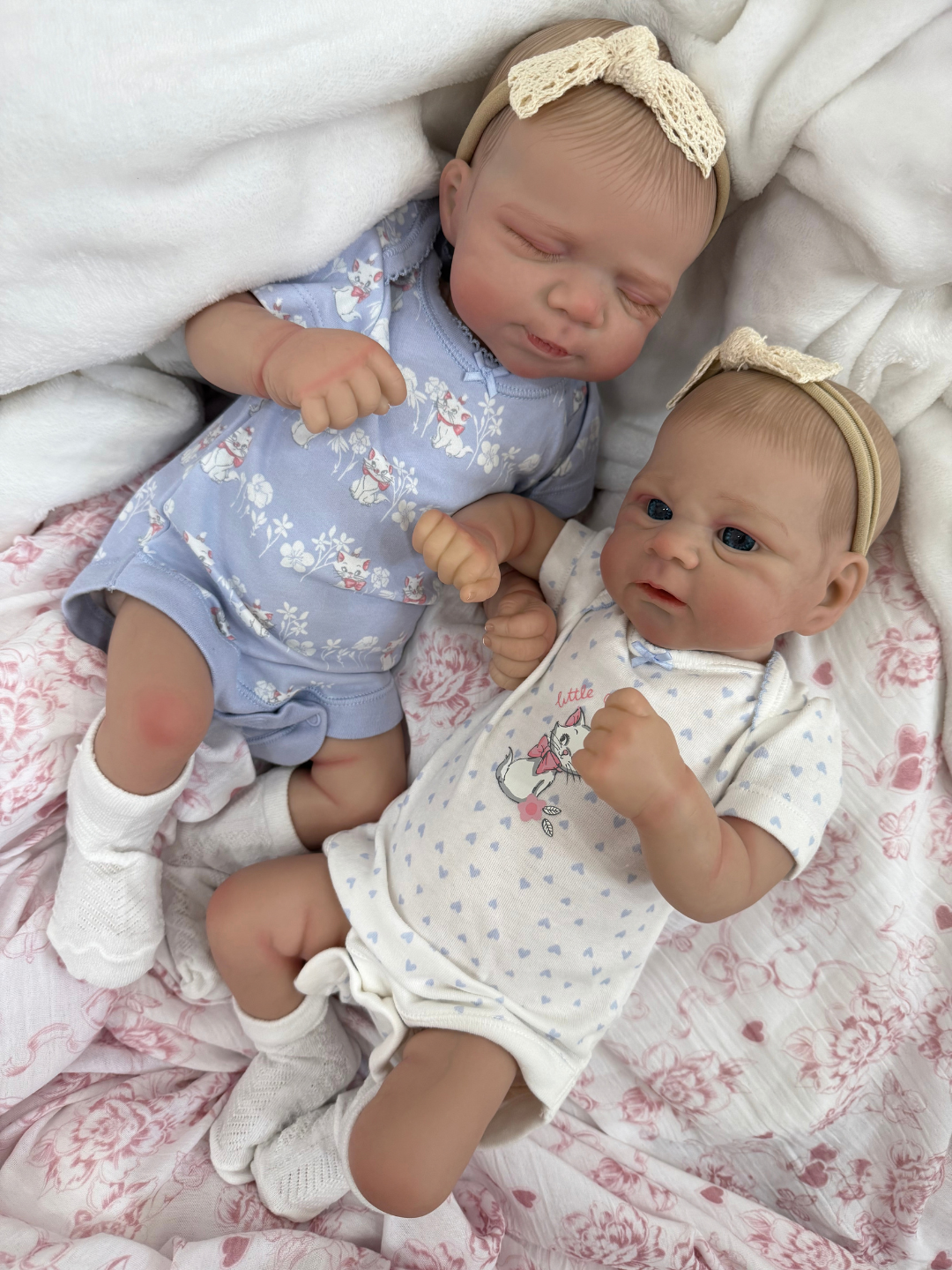 Two realistic baby dolls lying side by side on a pink floral blanket with white towels. One doll wears a light blue floral onesie and white socks, and the other wears a white heart-patterned onesie with a small pink flower design.