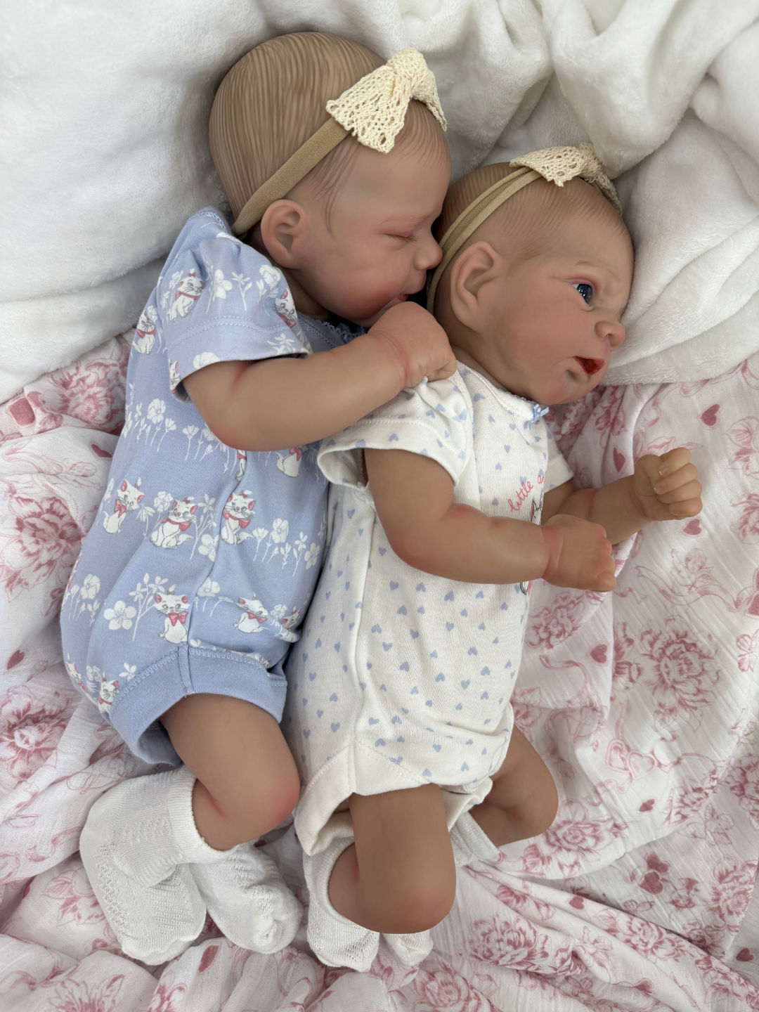 Two realistic baby doll twins lying side by side on a pink floral blanket, wearing matching beige lace headbands and one light blue and one white patterned onesie.
