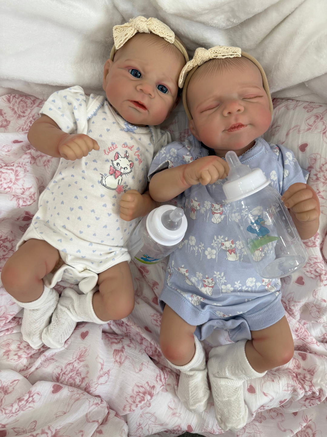 Two realistic reborn baby dolls lying side by side on a pink and white patterned blanket. The doll on the left wears a white onesie with a blue bunny pattern and a cream bow headband, while the doll on the right wears a light blue floral onesie and holds a clear bottle.