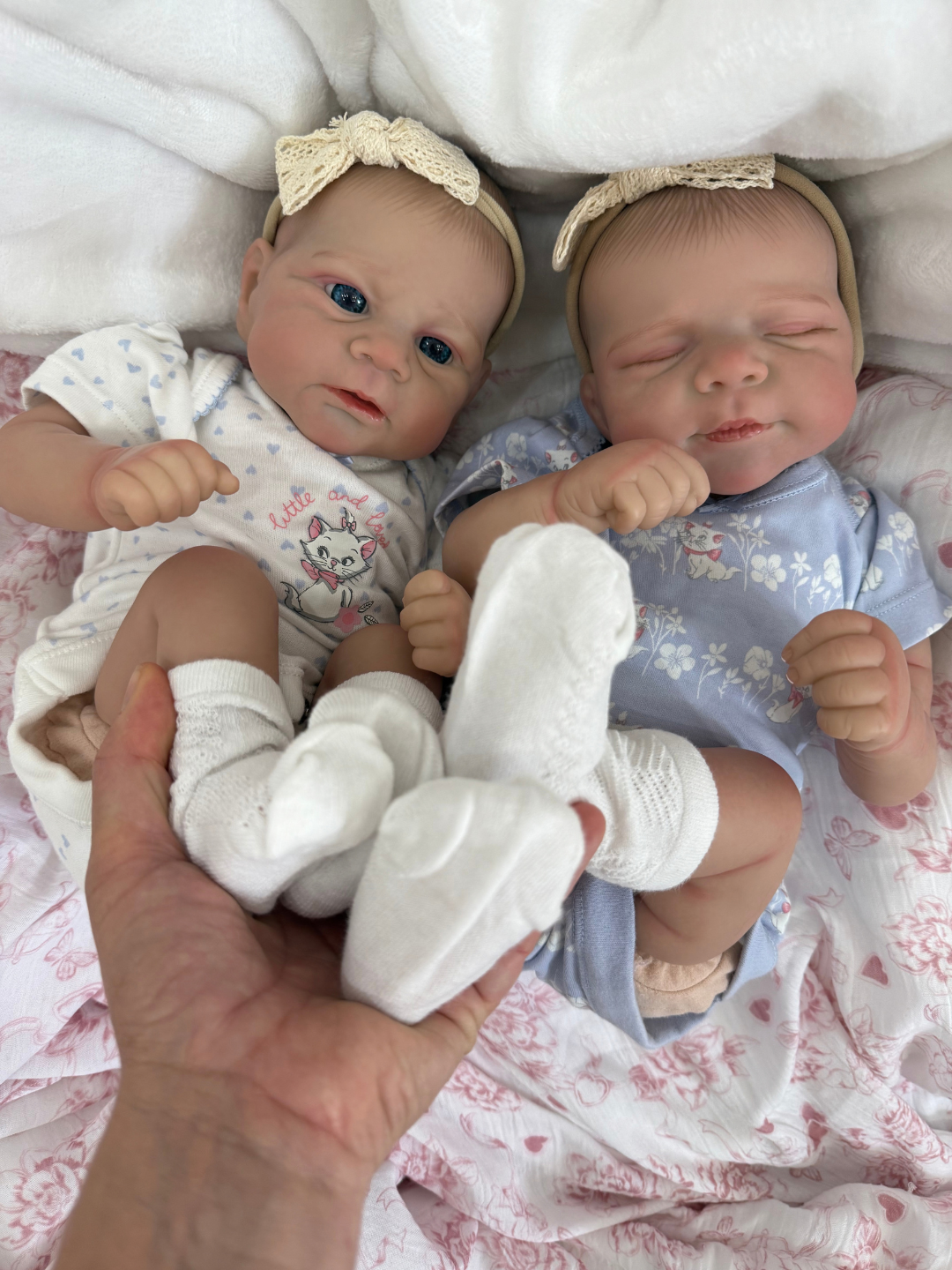 Two realistic reborn baby dolls lying side by side on a pink floral blanket, wearing matching outfits and white knit socks, with a human hand holding one doll's feet.