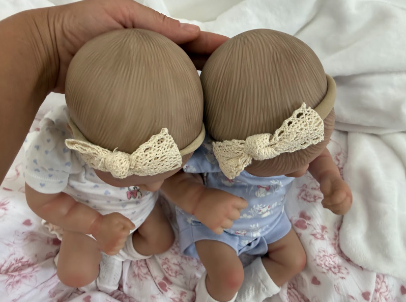 Two realistic baby dolls viewed from the back, each with textured light brown hair, cream lace bows, and matching outfits, held by a hand over white bedding.