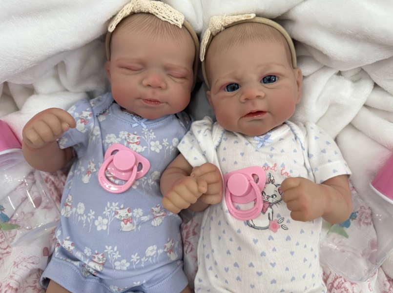 Two realistic baby doll twins lying side by side on a white blanket, wearing floral and heart-patterned bodysuits, matching beige headbands, and pink pacifiers.